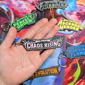 May include: A black and light pink keychain featuring the text Mega Evolution Chaos Rising in a stylized font. The keychain has a textured surface and is attached to a thin pink cord. Other similar keychains are visible in the background.