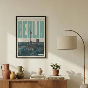 Könnte beinhalten: Ein gerahmtes Poster mit dem Wort "BERLIN" in großen, türkisfarbenen Buchstaben. Das Poster zeigt eine Stadtlandschaft mit dem Berliner Fernsehturm. Das Bild ist in einem schwarzen Rahmen und hängt an einer weißen Wand.