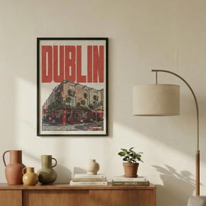 May include: Framed art print featuring a street scene in Dublin, Ireland, with the city name in large red letters at the top. The image shows a red pub with people and a street view. The print is displayed above a wooden cabinet.