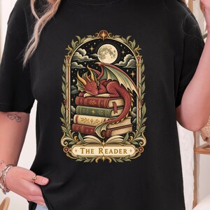 May include: A black t-shirt featuring a graphic print of a red dragon sleeping on a stack of books under a full moon. The design is framed by ornate foliage and includes the text "THE READER" at the bottom in a decorative banner style.