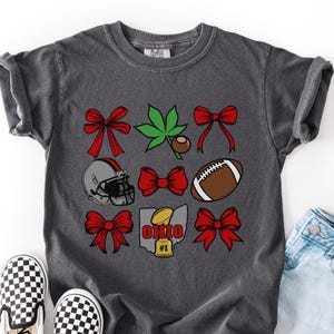 May include: Dark gray t-shirt featuring a grid of Ohio-themed graphics. The design includes red bows, a green leaf, a football helmet, a football, and a map of Ohio with a football and the text "OHIO #1".