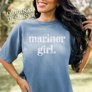 May include: A blue t-shirt with the words "mariner girl" printed in white. The shirt has short sleeves and a crew neck. The words "ORIGINAL DESIGNER" are printed in white at the top left.