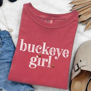 May include: A red t-shirt with the words "buckeye girl" in white script. The shirt has a crew neck and short sleeves. Two small brown football graphics are below the word "girl".