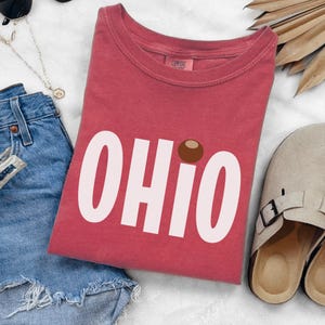 May include: A red t-shirt with the word "OHIO" in large white letters. The "O" is replaced with a brown circle. The shirt is folded and placed on a white surface, with denim shorts and sandals nearby.