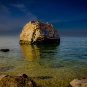 Puede incluir: Una gran isla rocosa se encuentra en medio de un océano azul tranquilo. El agua es clara y poco profunda, revelando el fondo arenoso. El cielo es de un azul profundo con algunas nubes blancas.