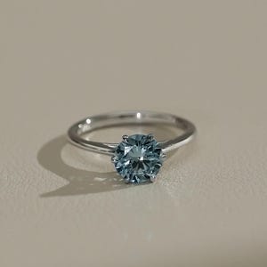 Peut inclure: Une bague argentée ornée d'une pierre précieuse ronde à facettes de couleur bleu ardoise, fixée par un sertissage à six griffes. L'anneau est lisse et poli, présentant un design épuré qui met en valeur la teinte bleue froide et mate.
