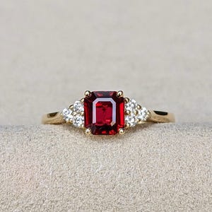 Könnte beinhalten: Ein Goldring mit einem zentralen, smaragdgeschliffenen, tiefroten Edelstein, der von vier Krappen gehalten wird. Der Ring ist auf jeder Seite mit einer Gruppe von drei kleinen, runden, klaren weißen Steinen im Goldband verziert.