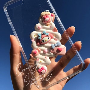 May include: A clear phone case with a 3D design of a white and pink swirl with a pink Hello Kitty character on top. The design includes pink and blue bows, hearts, and sprinkles. The text "Creamy Spring" is written on the case.