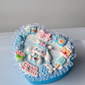 Könnte beinhalten: Eine herzförmige Schmuckschatulle mit blauem Boden und weißem Spitzensaum. Die Schatulle ist mit einer Zeichentrickfigur, Schleifen und Miniatur-Lebensmitteln verziert. Der Text "CINNA ROLL" ist sichtbar.