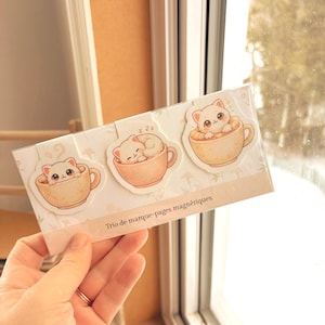 Peut inclure: Un ensemble de trois marque-pages magnétiques illustrant des chatons blancs dans des tasses à café beiges. L'emballage est une carte rectangulaire avec le texte Trio de marque-pages magnétiques imprimé en brun foncé sur la partie inférieure.