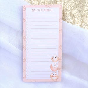 Peut inclure: Un bloc-notes rose avec le texte "MA LISTE DU MOMENT" en haut. Le bloc-notes présente un format de liste avec des cases à cocher et des illustrations de chatons dans des tasses à café. Des illustrations de grains de café sont sur le côté gauche.