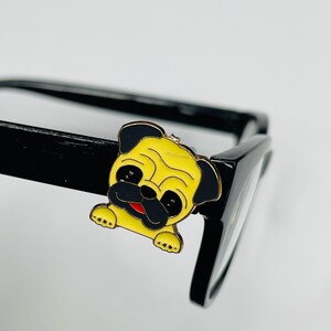 May include: A yellow and black pug dog charm attached to a pair of black spectacles. The charm has a red mouth and is made of metal. The spectacles have a thick frame.