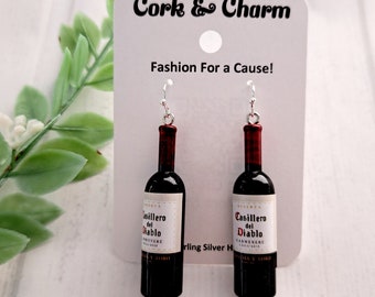 May include: A pair of silver earrings featuring miniature wine bottles with the label "Casillero del Diablo" printed on them. The bottles are red and have a silver cork.