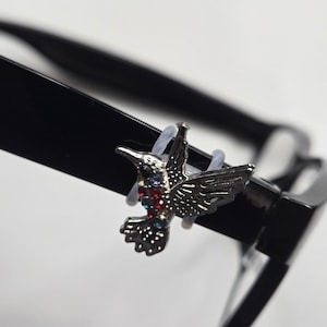 May include: A silver-toned hummingbird accessory attached to the black temple of a pair of eyeglasses. The hummingbird features small embedded red, blue, and black rhinestones along its body and wings, secured by a clear, flexible plastic loop.