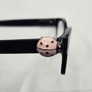 May include: A black-framed pair of eyeglasses with a pink ladybug-shaped decorative clip attached to the frame. The ladybug has black spots and gold accents. The background is a plain white.