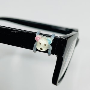Puede incluir: Gafas negras con un accesorio decorativo transparente en forma de oso sujeto a la montura. El oso tiene detalles en rosa y azul y ojos negros en forma de cruz. Las gafas están sobre un fondo blanco.