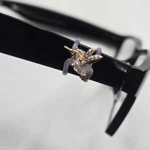 May include: A gold-toned hummingbird-shaped accessory with clear, sparkling stones attached to the side of a black eyeglass frame. The hummingbird features a teardrop-shaped stone for the body and smaller stones along the wings and head.