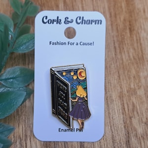 May include: Enamel pin featuring a book with a starry night sky and a girl in a purple dress. The pin is on a white card with the text "Cork & Charm" and "Fashion For a Cause!"