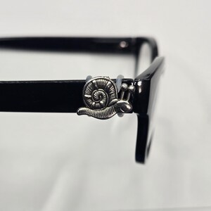 May include: Black eyeglasses with a silver snail charm attached to the frame. The snail charm is detailed with a spiral shell and antennae. The glasses have a rectangular shape and a glossy finish.
