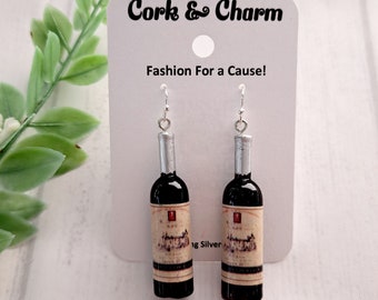 May include: A pair of silver dangle earrings featuring miniature wine bottles with cork tops and vintage labels. The earrings are displayed on a white card with the text "Cork & Charm" and "Fashion For a Cause!"