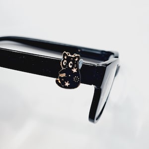 May include: A black cat-shaped eyeglass accessory with gold accents, featuring stars and a flower design. The accessory is attached to a pair of black eyeglasses.