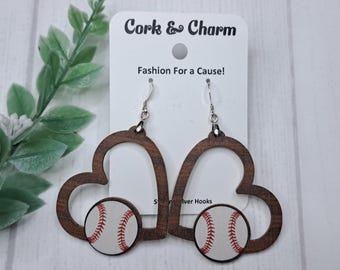 Puede incluir: Un par de pendientes de madera en forma de corazón con un diseño de béisbol. Los pendientes son marrones con una pelota de béisbol blanca y costuras rojas. Los pendientes tienen ganchos plateados.