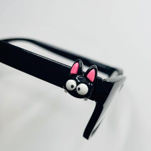 Puede incluir: Gafas negras con una cara de gato negro decorativa en la montura. La cara del gato tiene orejas internas rosas y ojos grandes y blancos. Las gafas están en ángulo, mostrando la montura y parte de la lente.