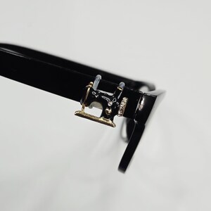 May include: Black eyeglasses with a decorative sewing machine charm on the frame. The charm is gold-toned with black enamel and rhinestone accents. The glasses have a sleek, modern design.