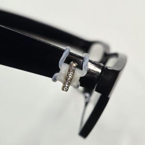 May include: Close-up of black eyeglasses with a decorative, gold-colored bar adorned with small, clear rhinestones. The bar is attached to the glasses with clear, flexible loops. The glasses are set against a white background.
