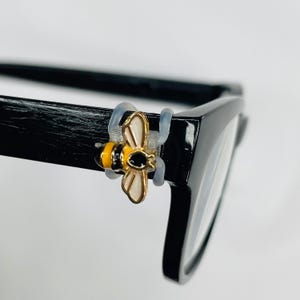 May include: A black eyeglass frame with a decorative bee charm attached. The bee charm is yellow, black, and gold, and is secured to the frame with clear silicone loops. The glasses have a rectangular shape.