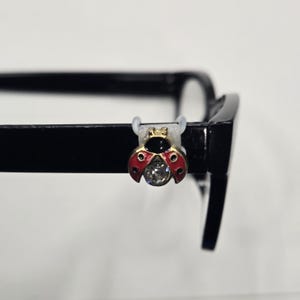 May include: A decorative ladybug eyeglass charm. The charm is red with black spots and a gold-colored head. A clear, faceted gem is in the center. The charm is attached to the black frame of a pair of eyeglasses with a clear plastic loop.