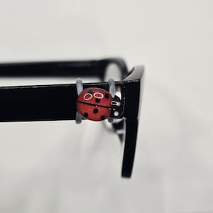 May include: A pair of black eyeglasses with a red ladybug charm attached to the frame. The ladybug charm is red with black spots and white eyes. The glasses are set against a white background.