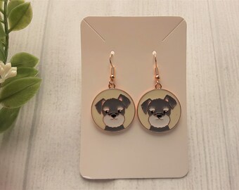 Puede incluir: Un par de pendientes colgantes de tono dorado con la cara de un perro schnauzer miniatura en blanco y negro sobre un fondo crema.