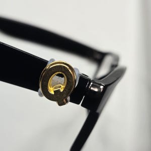 May include: Close-up of black eyeglasses with a gold-colored letter "Q" emblem on the frame. The glasses have a classic design with a thick, rectangular frame and a shiny finish. The emblem is circular and attached to the frame.