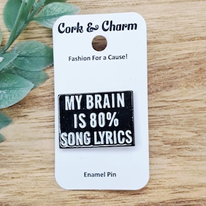 Könnte beinhalten: Ein schwarzer Emaille-Pin mit dem weißen Text "MY BRAIN IS 80% SONG LYRICS". Der Pin befindet sich auf einer weißen Karte mit dem Text "Cork & Charm" und "Fashion For a Cause!". Auf der Karte steht auch "Enamel Pin".
