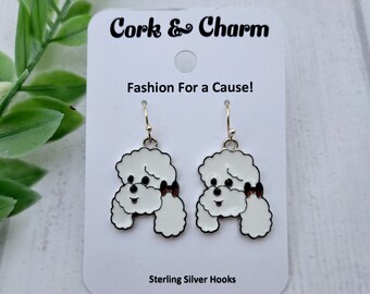 Puede incluir: Pendientes colgantes de perro caniche blanco con un lazo rojo. Los pendientes están en una tarjeta blanca con el texto "Cork & Charm Fashion For a Cause! Sterling Silver Hooks".