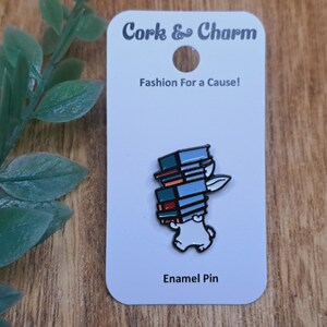 Pode incluir: Pin de esmalte branco com um coelho de desenho animado carregando uma pilha de livros coloridos. O pin diz "Cork & Charm" e "Fashion For a Cause!" e "Enamel Pin".