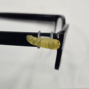 May include: A close-up of a pair of black eyeglasses with a yellow caterpillar-shaped accessory attached to the frame. The caterpillar has two small black dots for eyes and is made of a translucent material.
