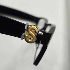 May include: Close-up of black eyeglasses with a gold-colored letter "S" emblem on the frame. The glasses have a classic rectangular shape and a glossy finish. The emblem is attached to the frame with a small, light-colored ring.