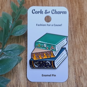 May include: A stack of three enamel pins depicting books with different designs. The top book is green with floral details, the middle book is brown with orange flames, and the bottom book is blue with stars and moons. The pins are on a white card with the text "Cork & Charm" and "Fashion For a Cause!" and "Enamel Pin".