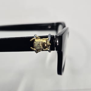 May include: Black eyeglasses with a gold-colored beetle charm on the left temple. The glasses have a rectangular frame and a glossy finish. The beetle charm is detailed with intricate design.