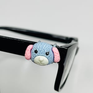 Puede incluir: Un accesorio para gafas con forma de perro azul, rosa y blanco, sujeto a una montura de gafas negra. El perro tiene una cabeza azul texturizada, orejas rosas y un hocico blanco. Las gafas son negras y brillantes.