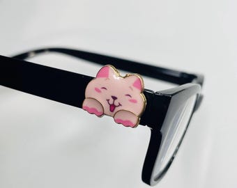 Op de afbeelding: Zwarte bril met een roze en gouden kattenbedel. De bedel heeft een lachend kattengezicht en roze pootjes. De bril heeft heldere glazen en een rechthoekige vorm.