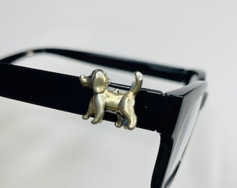 May include: A silver-colored dog charm attached to the black frame of a pair of eyeglasses. The dog is in a standing position with its tail up. The glasses have a rectangular shape.