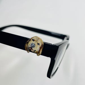 Puede incluir: Gafas negras con un adorno decorativo de perro golden retriever sujeto a la montura. El adorno es un retrato en miniatura detallado de la cara de un golden retriever, con colores marrón, beige y blanco. Las gafas tienen un acabado brillante.