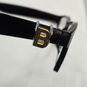 May include: Close-up of black eyeglasses with a gold-colored letter "B" on the left arm. The letter is attached with a clear plastic ring. The glasses have a thick frame and a modern design.