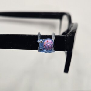 May include: A blue snail eyeglass accessory with a pink and purple shell is attached to a black eyeglass frame. The snail has a whimsical design, with a smiling face and a spiral shell pattern. The accessory is made of a glossy material.