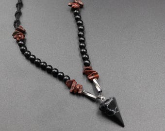 May include: A black beaded necklace with a pendant. The necklace features black beads, interspersed with reddish-brown stone chips. The pendant is a black, pointed shape with a white pattern, suspended by silver-colored metal.