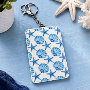 May include: A rectangular card holder featuring a white background with a repeating pattern of blue starfish and seashells. The card holder is attached to a silver-toned metal key ring with a short chain, allowing it to function as a keychain.