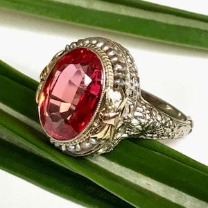 May include: A vintage-style ring featuring a large, oval-cut, ruby-red gemstone. The ring has an ornate, silver-toned band with intricate floral and filigree detailing. The gemstone is set in a gold-toned bezel, adding a touch of contrast and elegance.